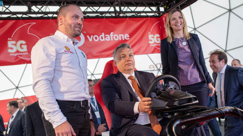 Orbán Viktor kormányfő a Vodafone autóját vezérli távvezérléssel, egy szimulátorral. A háttérben Mészáros Lőrinc is felsejlik